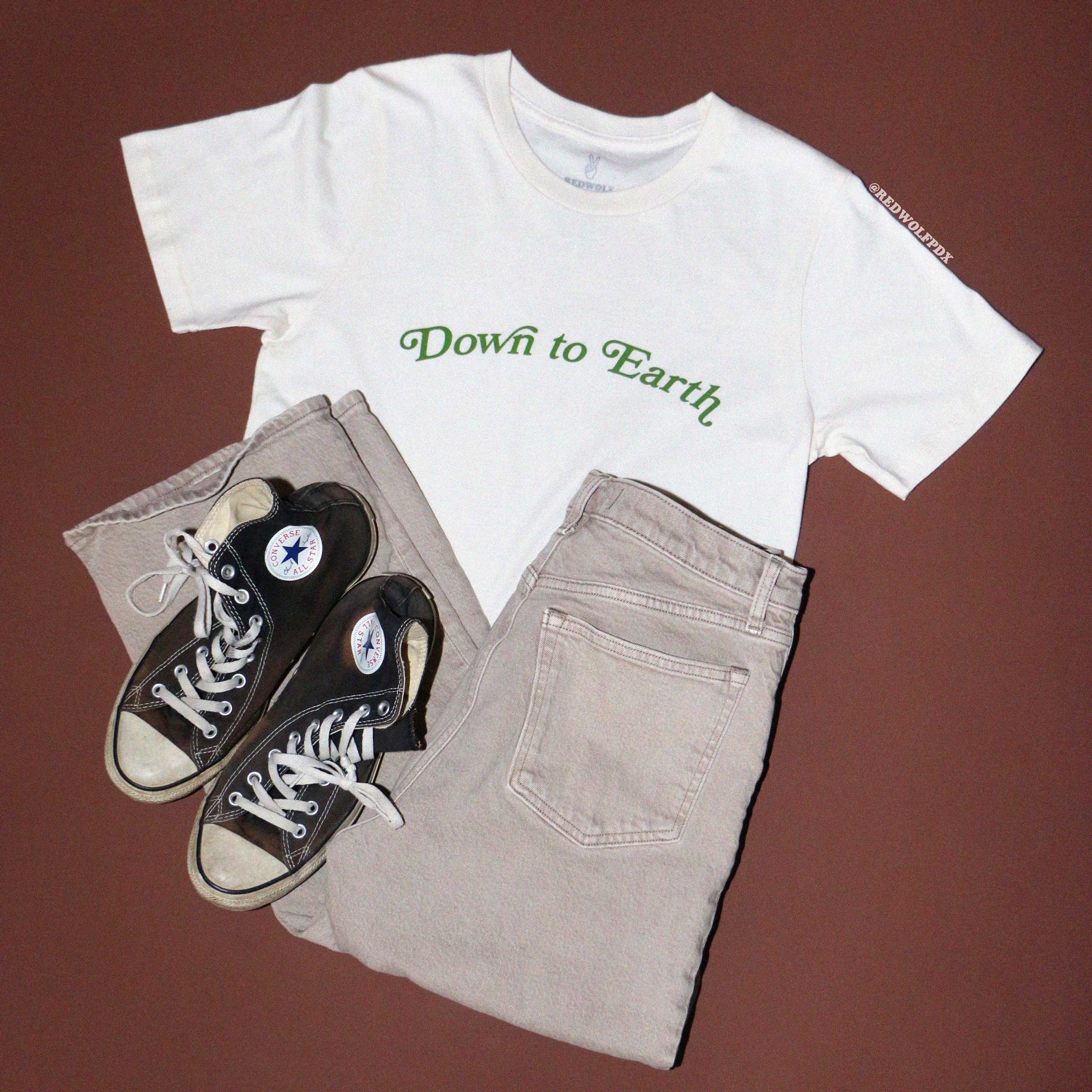 Graphic tee with text 'Down to Earth' with khaki jeans and sneakers for flatlay against brown backdrop