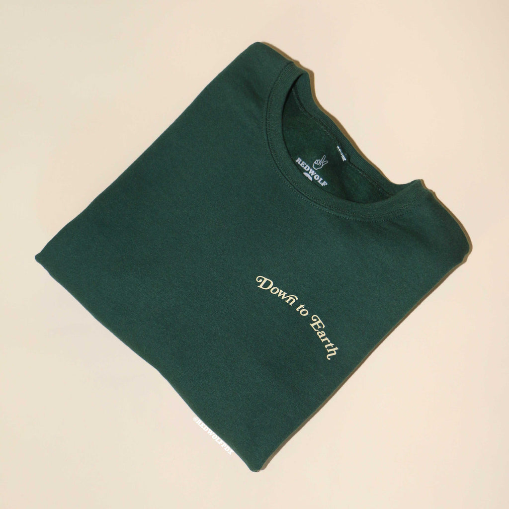 folded forest green crewneck sweatshirt with text 'Down to Earth' screen printed on front corner of chest on a beige background