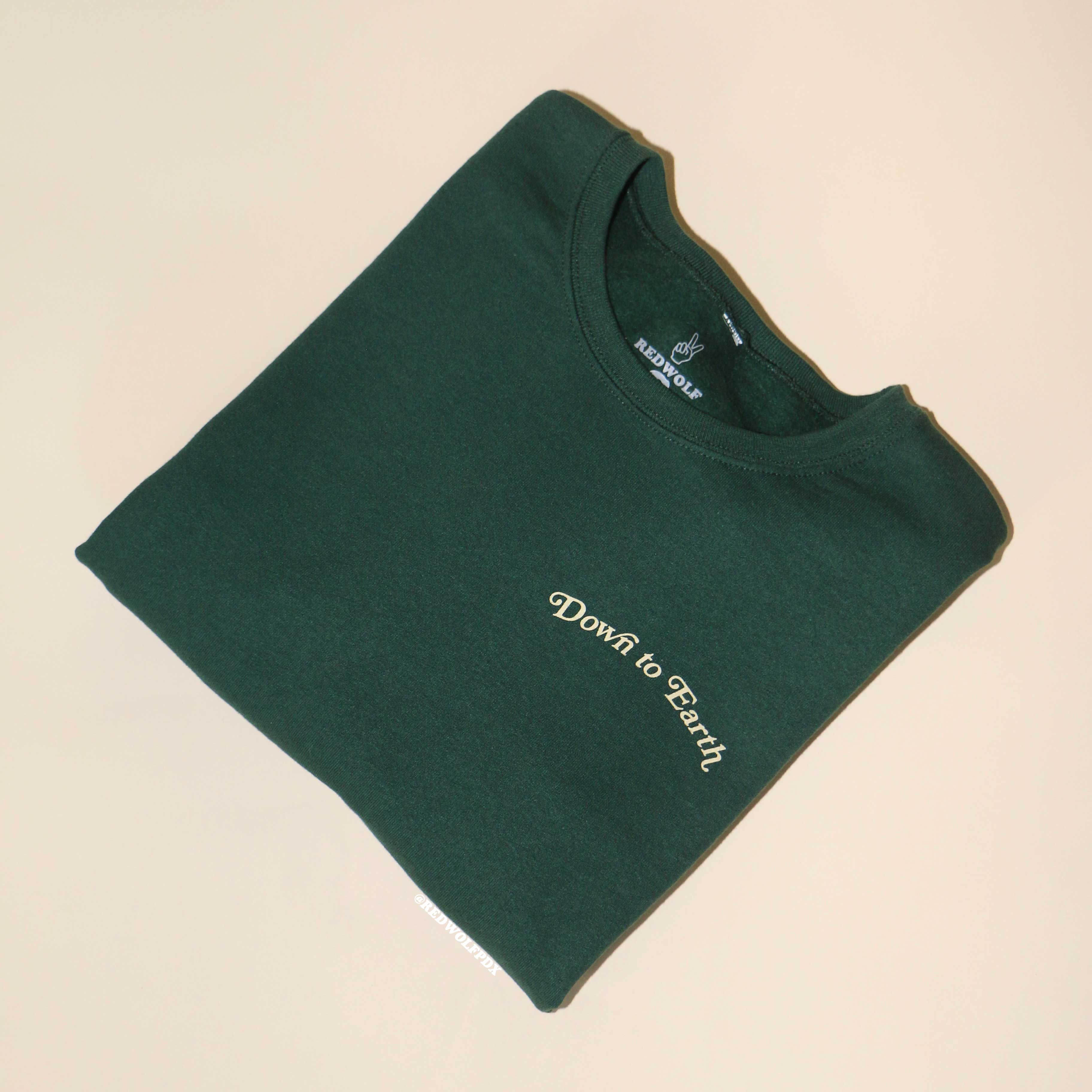 folded forest green crewneck sweatshirt with text 'Down to Earth' screen printed on front corner of chest on a beige background