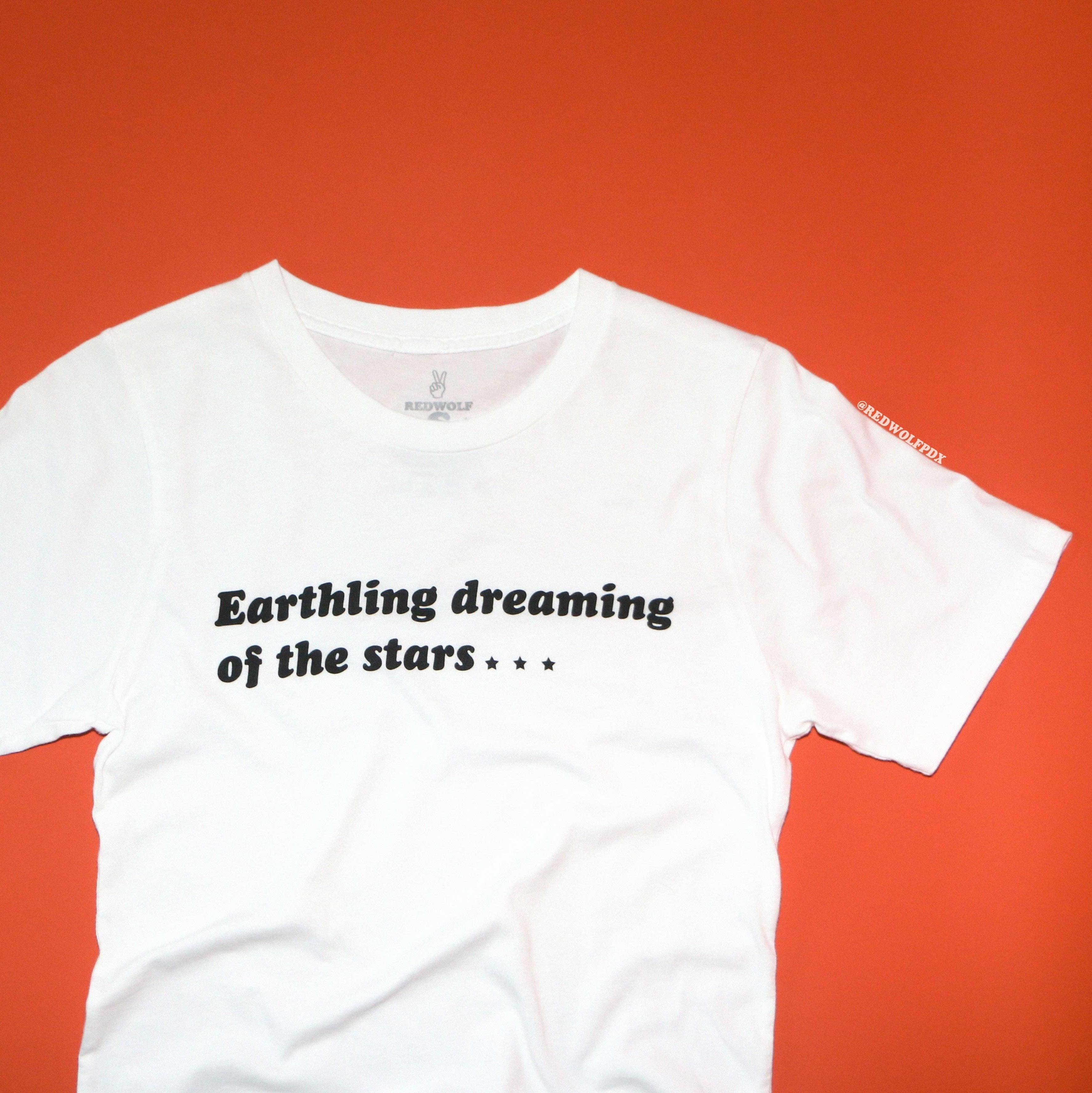 White t-shirt with text 'Earthling dreaming of the stars...' on an orange background