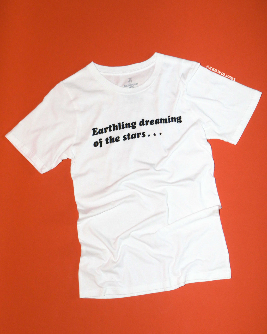 White t-shirt with text 'Earthling dreaming of the stars...' on an orange background