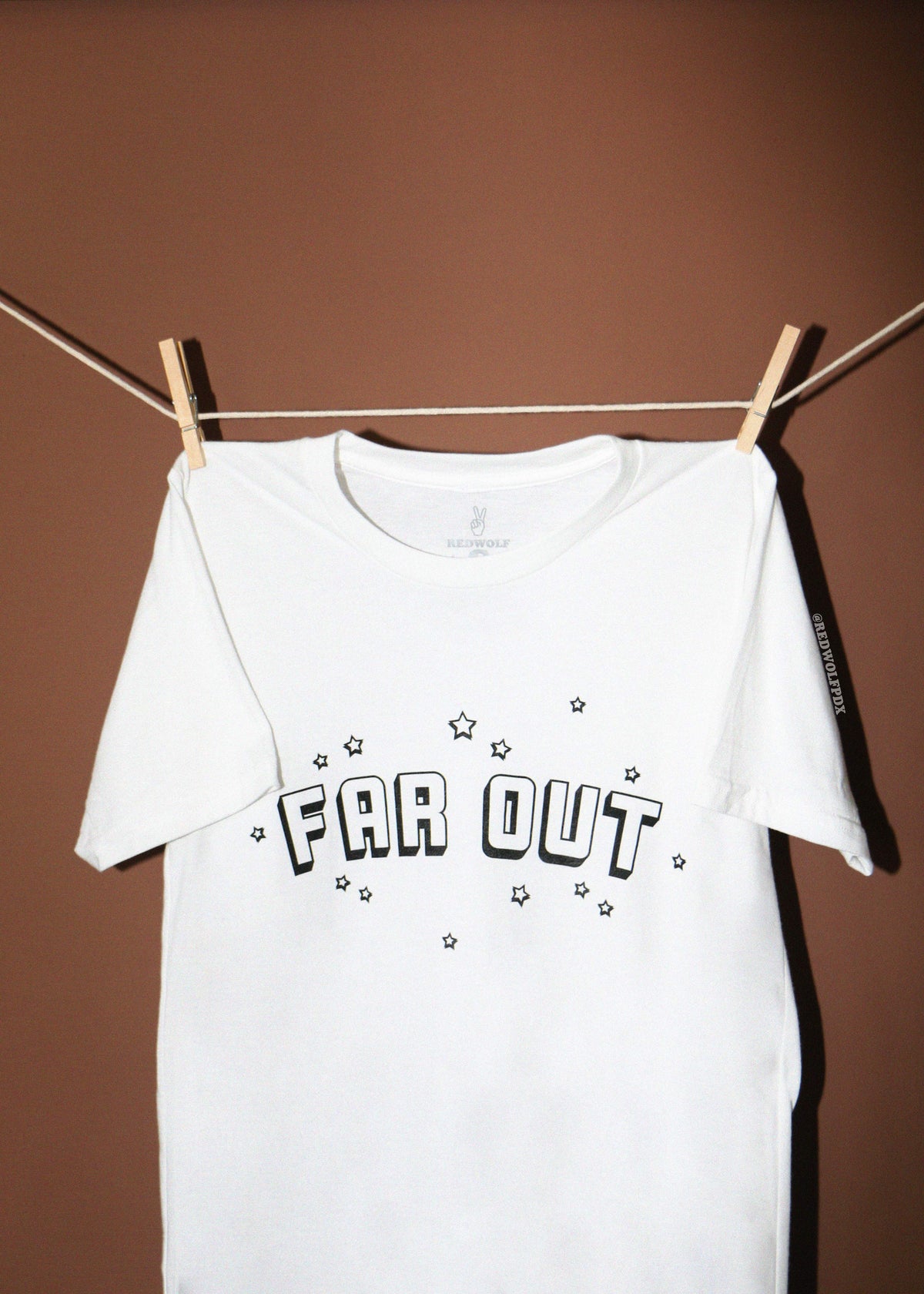 A white short-sleeve t-shirt with 'FAR OUT' and stars printed in black on the front, hanging from a clothesline with wooden clothespins against a solid brown background.