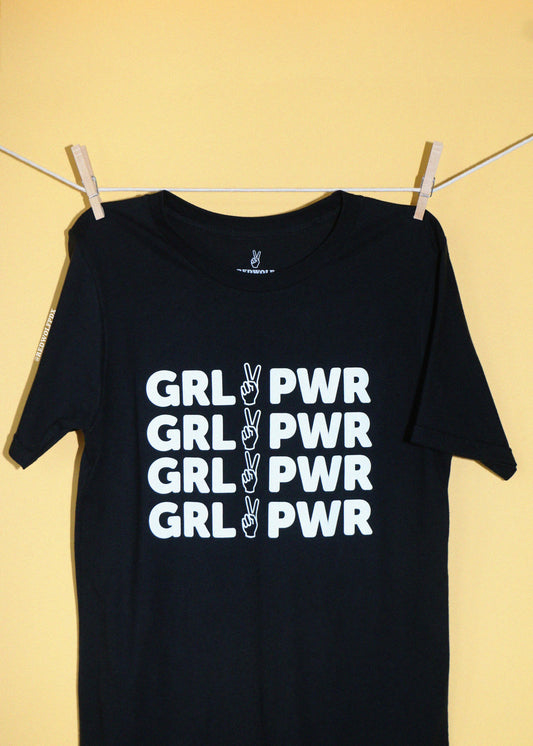 A black short-sleeve t-shirt hanging from a clothesline with wooden clips against a solid pale yellow background. The shirt features text 'GRL PWR' printed in white block letters, with a  a peace sign graphic, repeated four times vertically.