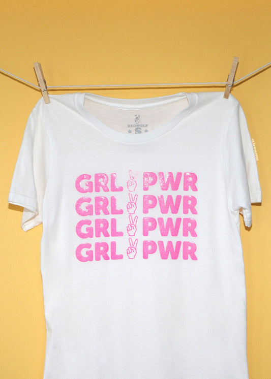 A white short-sleeve t-shirt hanging from a clothesline with wooden clothespins against a solid yellow background. The shirt features a graphic print of the text 'GRL PWR' repeated four times in pink glitter, with a peace sign graphic in the middle of each line.