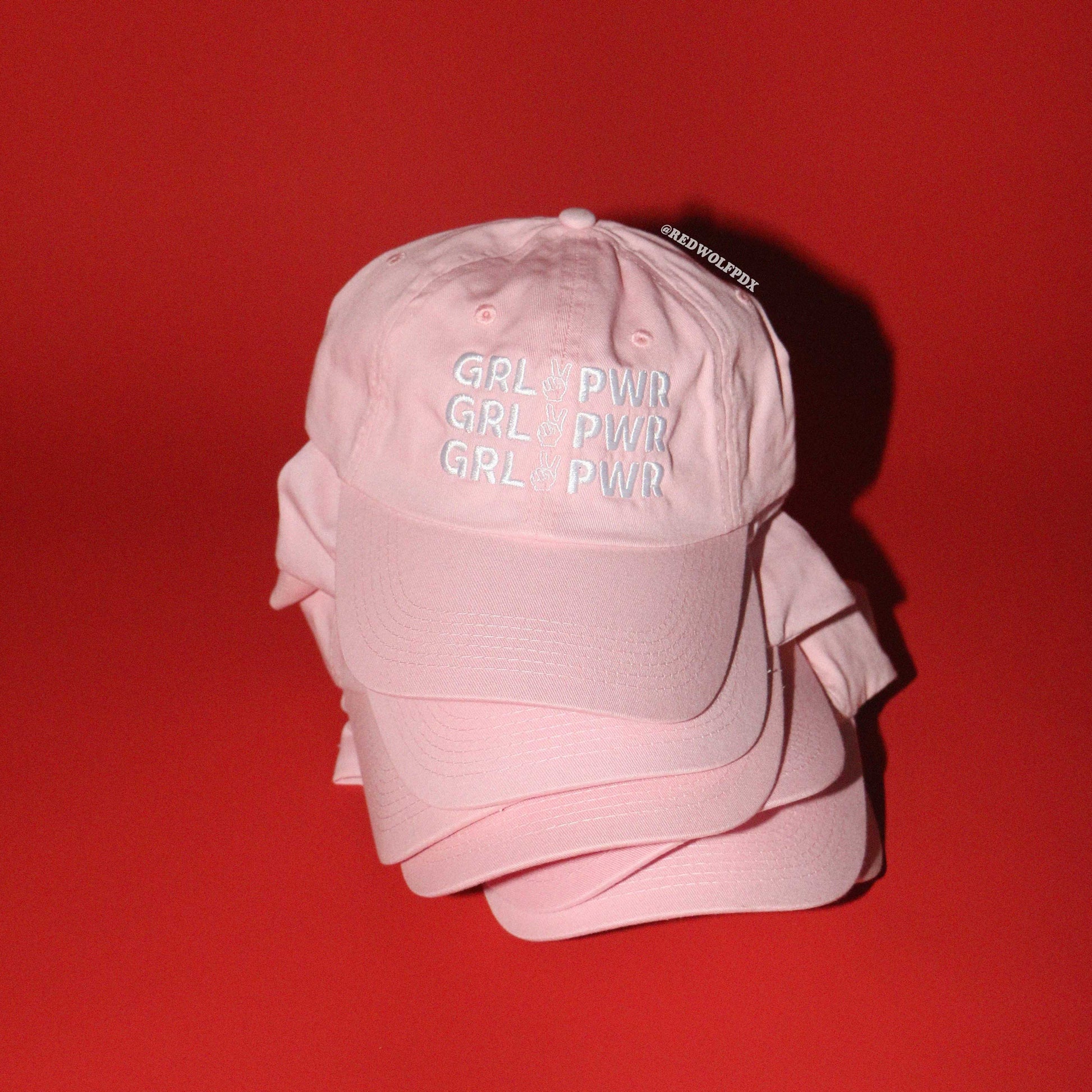 stack of light pink baseball caps with 'GRL PWR' text on a red background