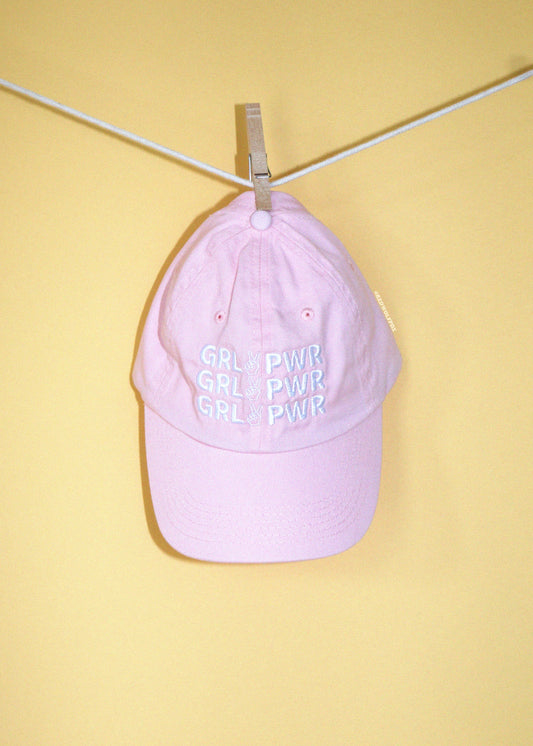 A light pink baseball cap with 'GRL PWR' embroidered in white thread on the front, hanging from a clothesline with a metal clip against a solid pale yellow background.