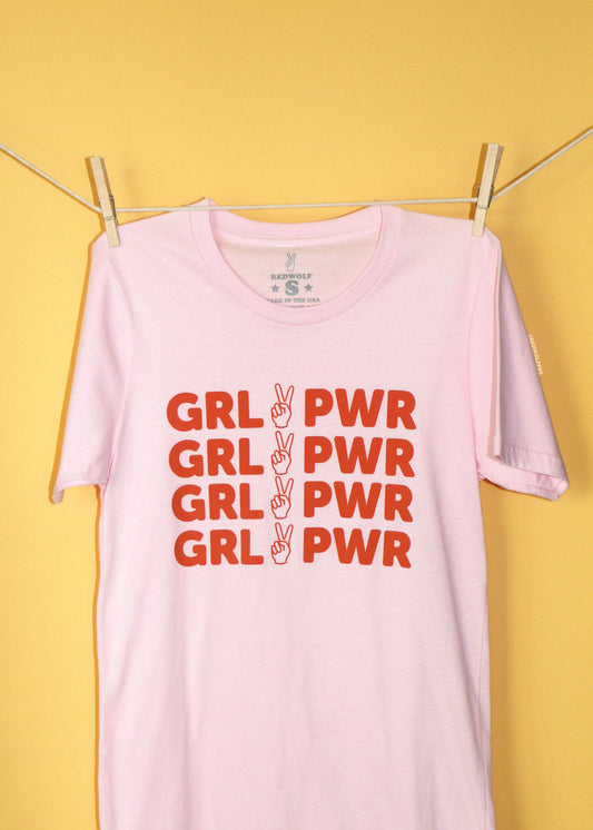 A light pink short-sleeve t-shirt hanging from a clothesline with wooden clothespins against a solid yellow background. The shirt features a graphic print in red text reading 'GRL PWR' repeated four times, with peace sign hand gesture graphic.