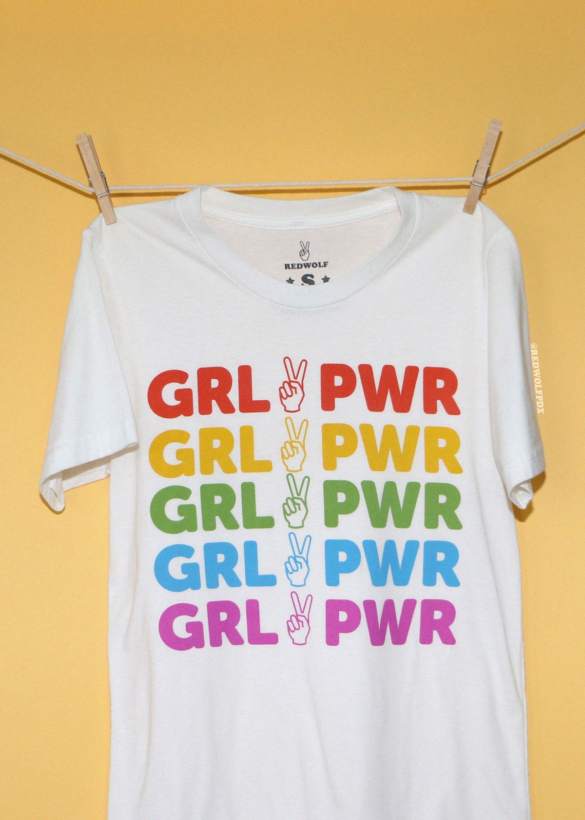 A white short-sleeve t-shirt hanging from a clothesline with wooden clips against a solid yellow background. The shirt features a graphic print of the text 'GRL PWR' repeated in red, yellow, green, blue, and pink, with a peace sign hand gesture in the center of each repetition.