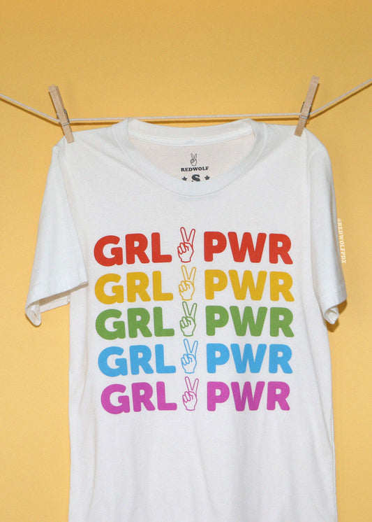 A white short-sleeve t-shirt hanging from a clothesline with wooden clips against a solid yellow background. The shirt features a graphic print of the text 'GRL PWR' repeated in red, yellow, green, blue, and pink, with a peace sign hand gesture in the center of each repetition.