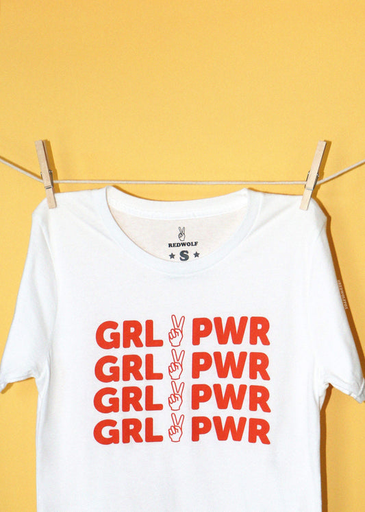 A white short-sleeve t-shirt with red text reading 'GRL PWR' repeated four times, hanging from a clothesline with wooden clips against a solid yellow background. The inner collar label shows the brand 'REDWOLF'.