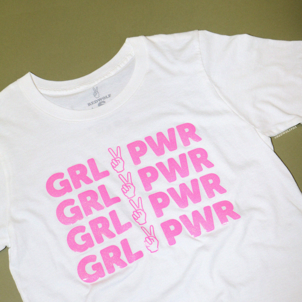 white tee with text 'GRL PWR' with peace sign graphic screen printed in pink glitter ink across the front against a vintage green background