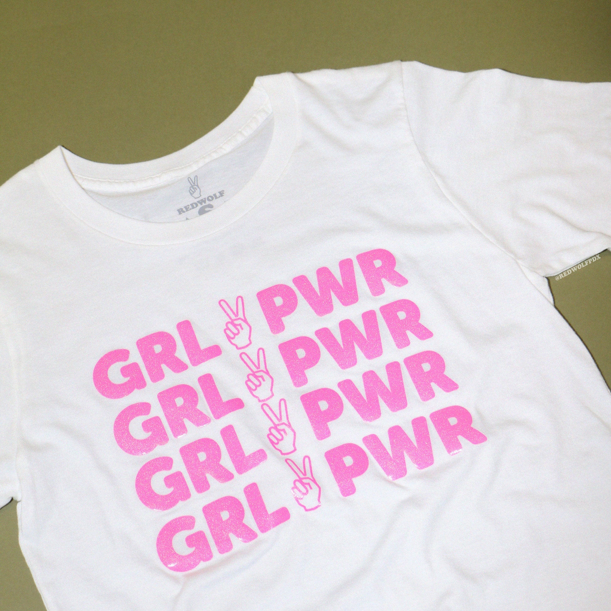 white tee with text 'GRL PWR' with peace sign graphic screen printed in pink glitter ink across the front against a vintage green background