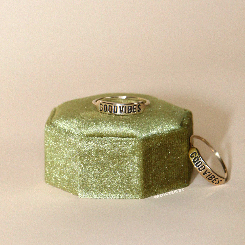 A ring with 'GOOD VIBES' engraved on it, displayed on a green textured stand.