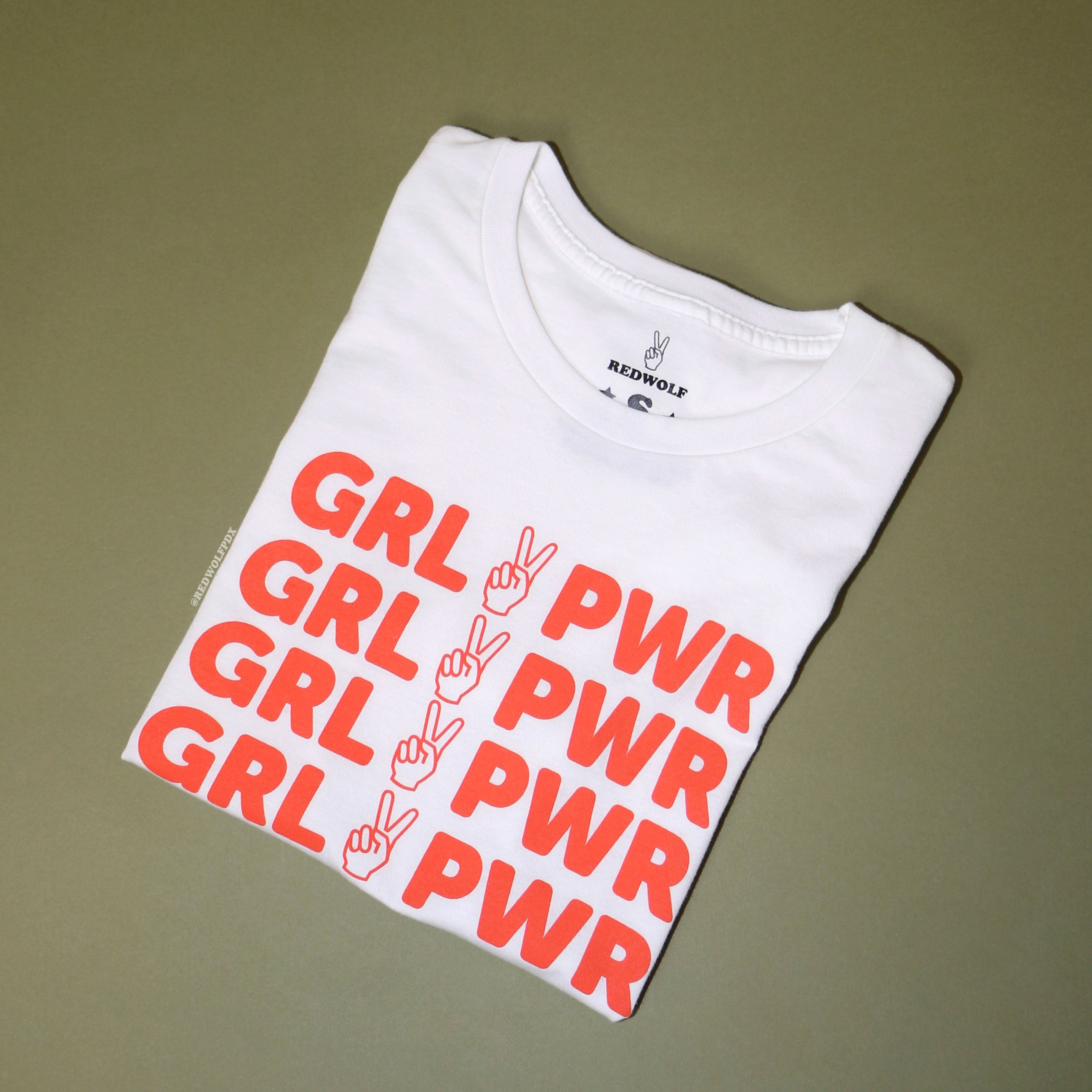 folded white graphic tee with text 'GRL PWR' and peace sign graphic in vintage red orange ink screen printed down the front against a vintage green background