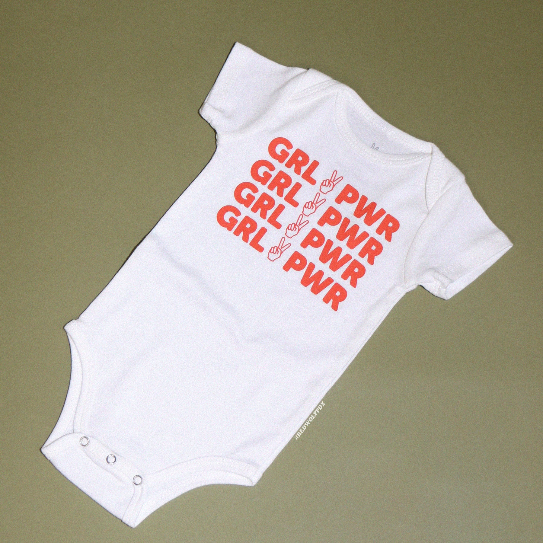 white onesie for babies with text 'GRL PWR' in orange red ink screen printed on front on a vintage green background
