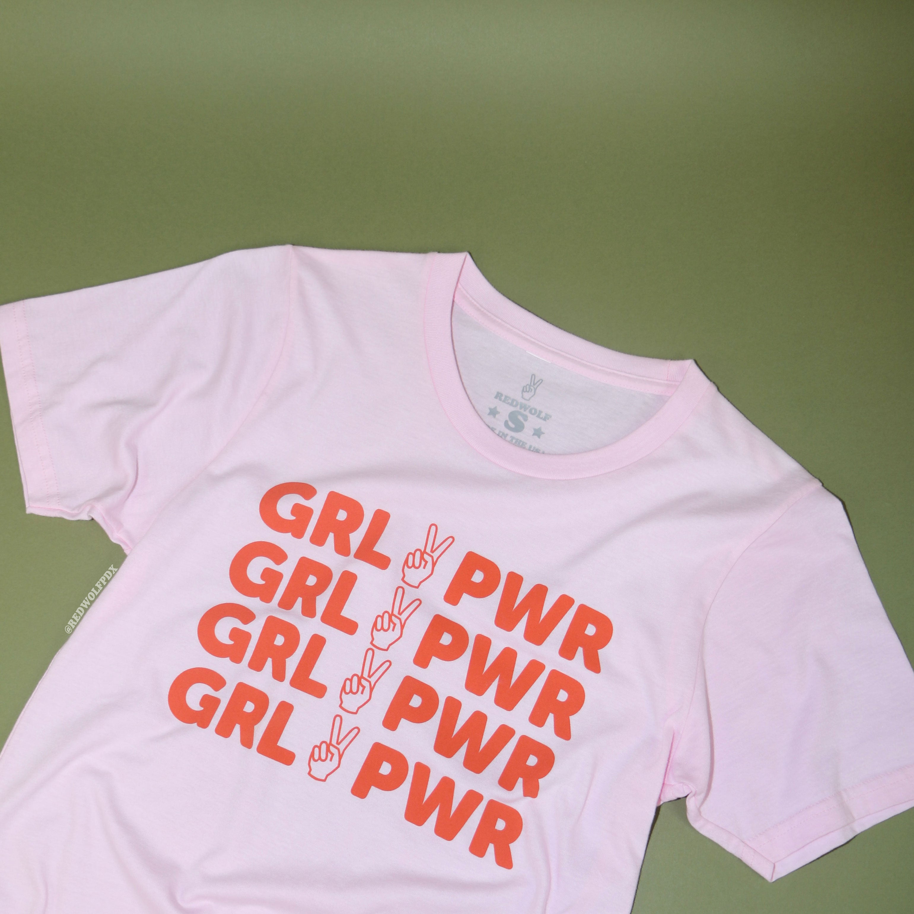 baby pink graphic tee with text 'GRL PWR' in orangey red ink screen printed on front against vintage green background