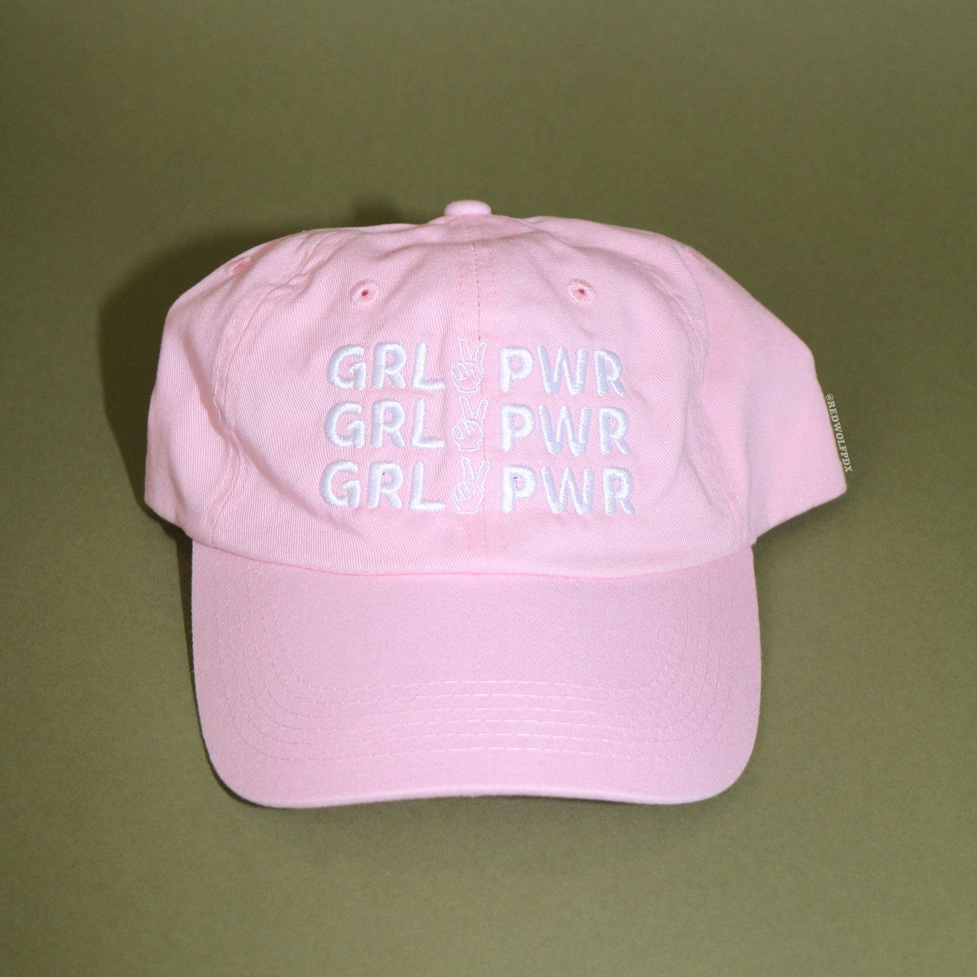 Pink baseball cap with text 'GRL PWR' embroidered in white thread across front on a green backdrop
