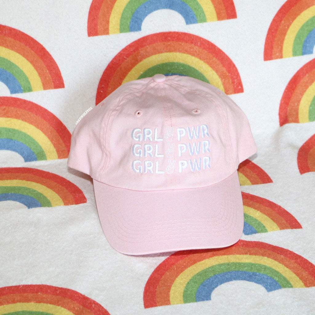 pink baseball cap with text 'GRL PWR' embroidered across front with white thread on a retro rainbow towel background
