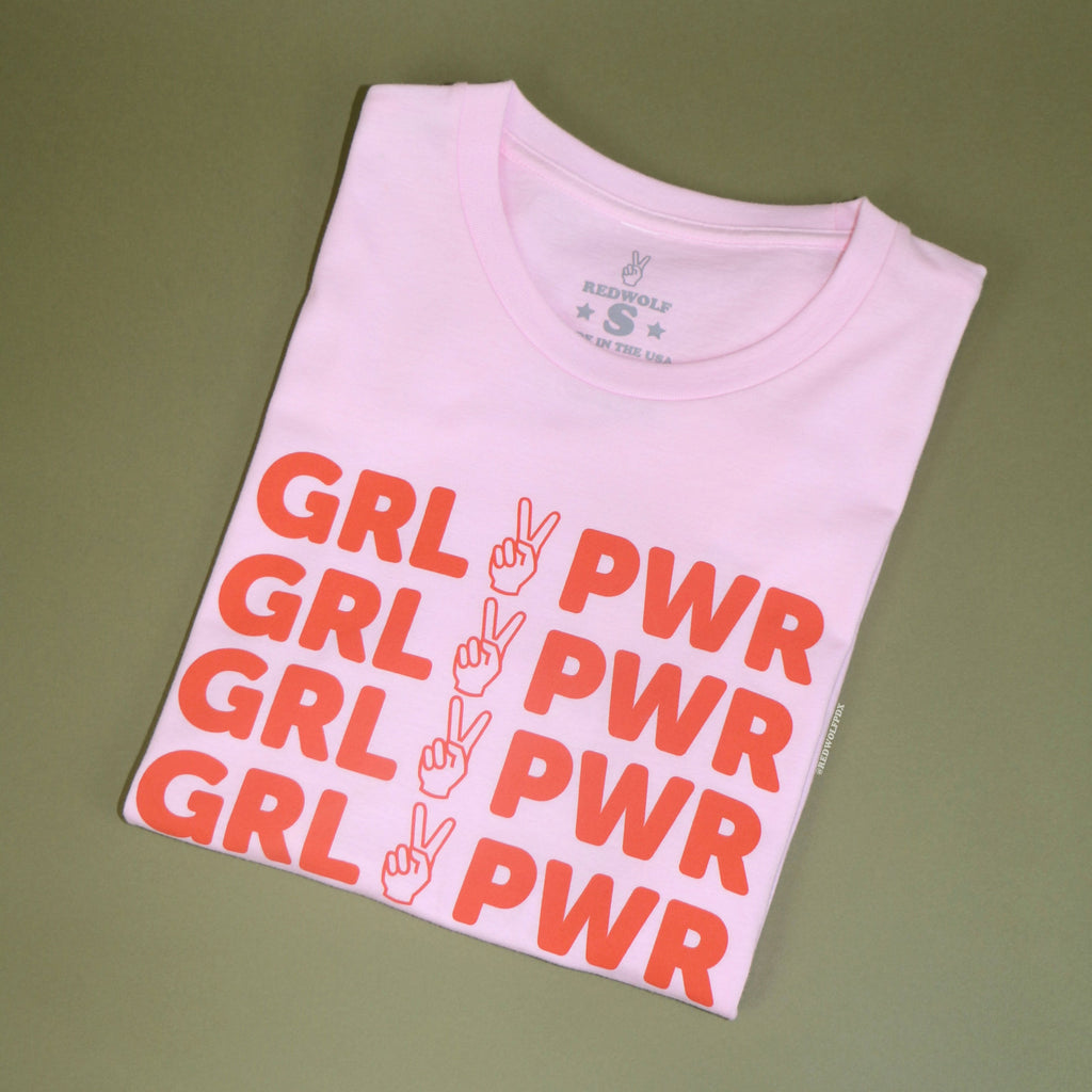 folded pink tee with text 'GRL PWR' with peace sign graphic in orange/red ink screen printed across chest against a vintage green background