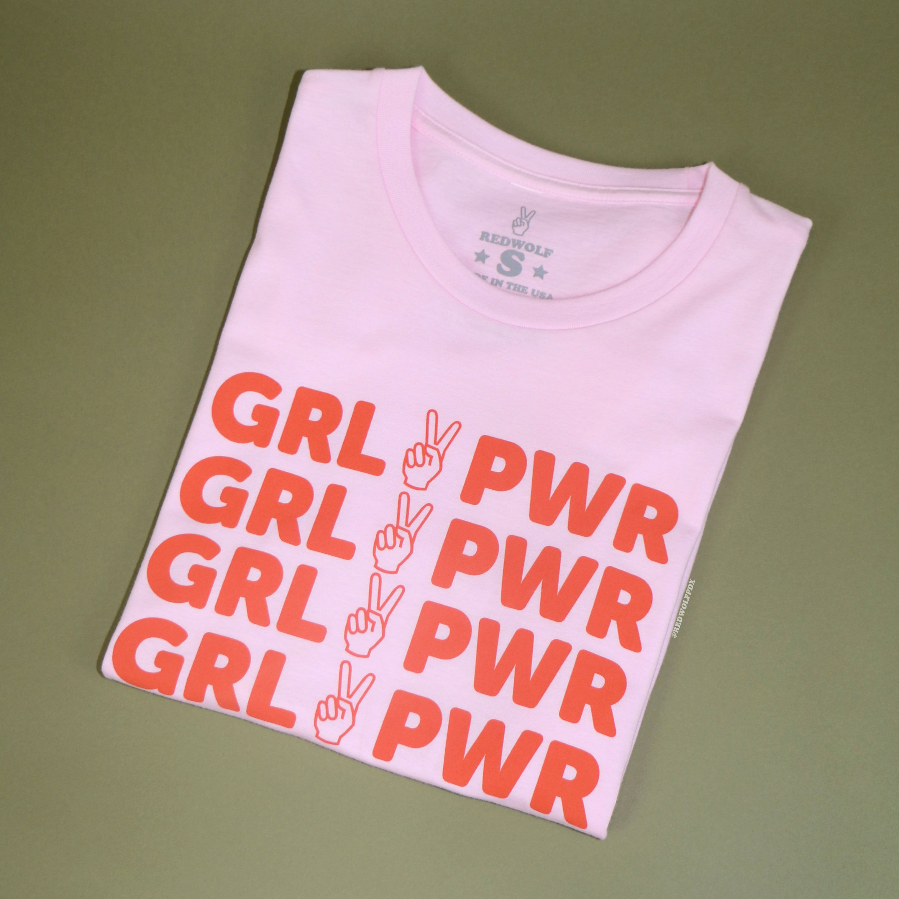 folded pink tee with text 'GRL PWR' with peace sign graphic in orange/red ink screen printed across chest against a vintage green background