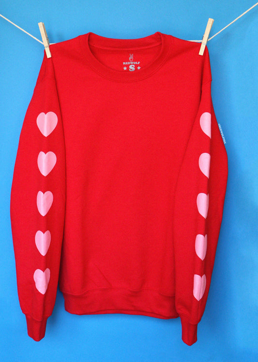 A red crewneck sweatshirt with pink heart graphics printed down both sleeves, hanging from a white clothesline with wooden clothespins against a bright blue background.