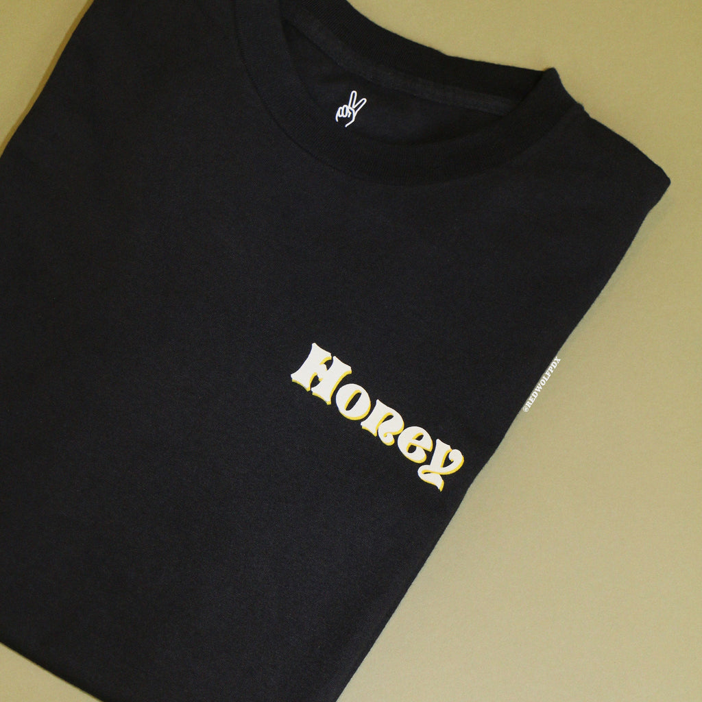 Folded black graphic tee with text 'Honey' in layered white and yellow ink screen printed across front corner against vintage green background.