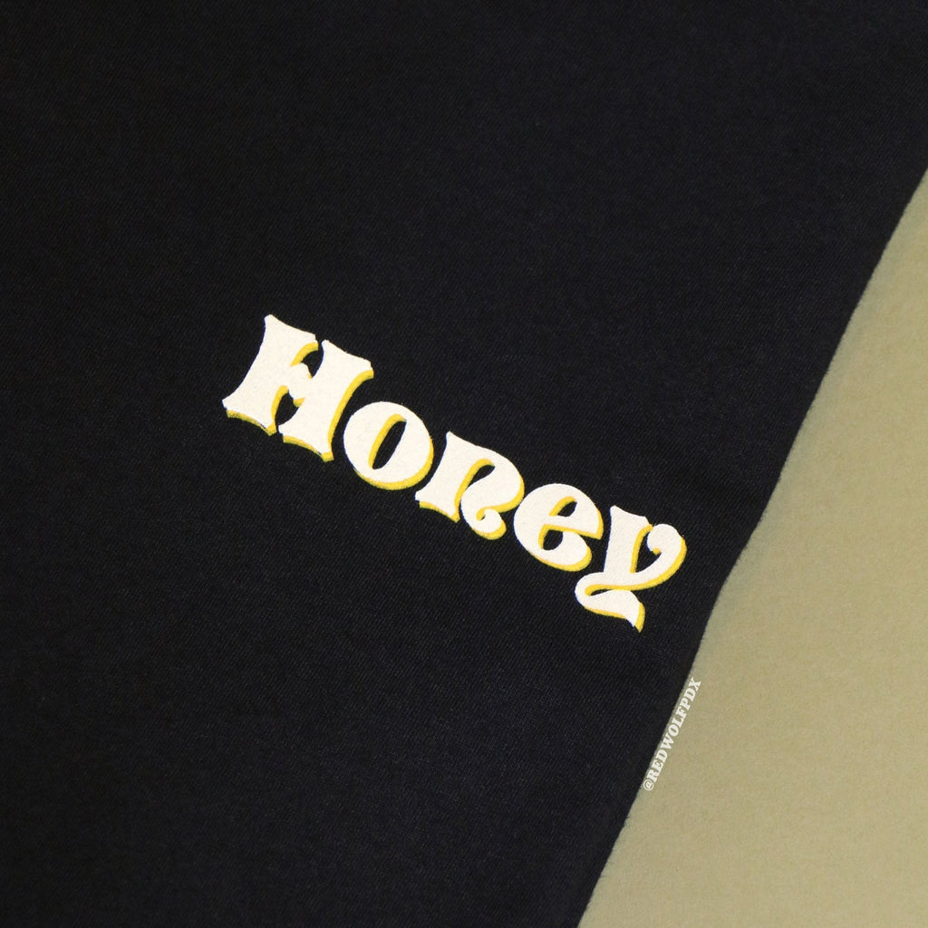 Up close shot of black graphic tee with text 'Honey' in white ink shadowed with yellow ink screen printed on front corner against vintage green background