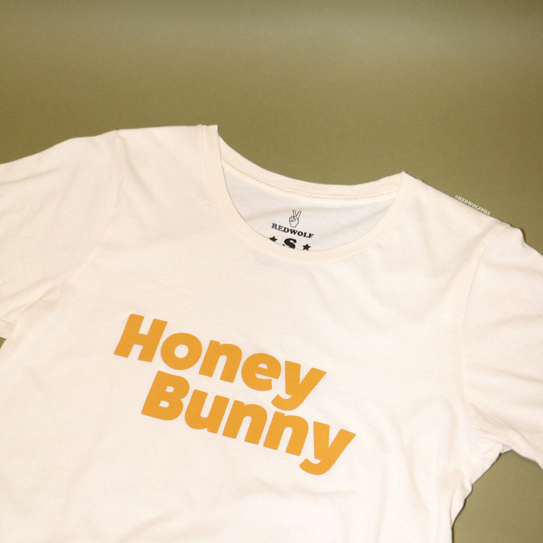 Beige graphic tee with text 'Honey Bunny' in vintage golden yellow ink screen printed across front against vintage green background