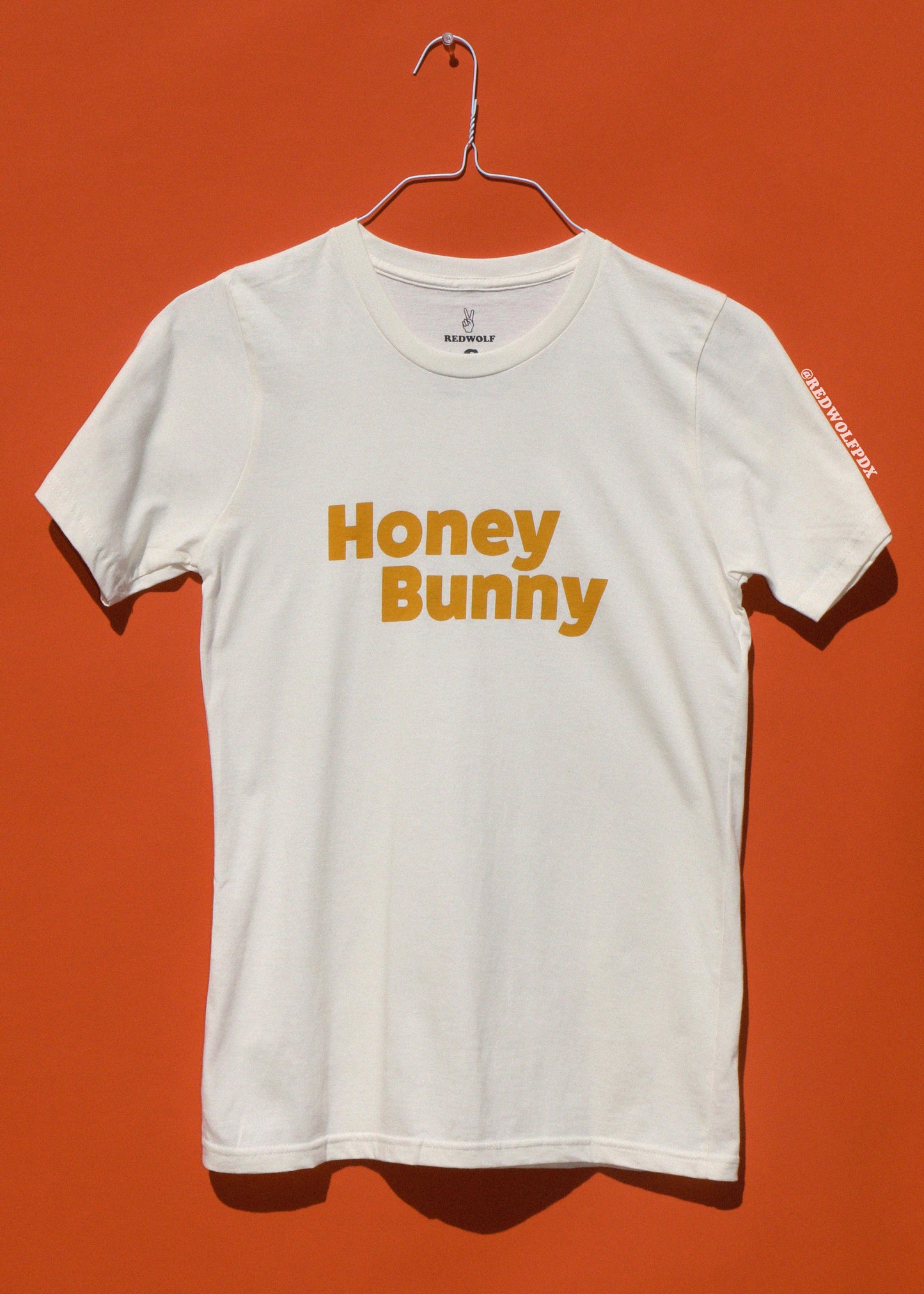 White t-shirt with 'Honey Bunny' text on a hanger against an orange background