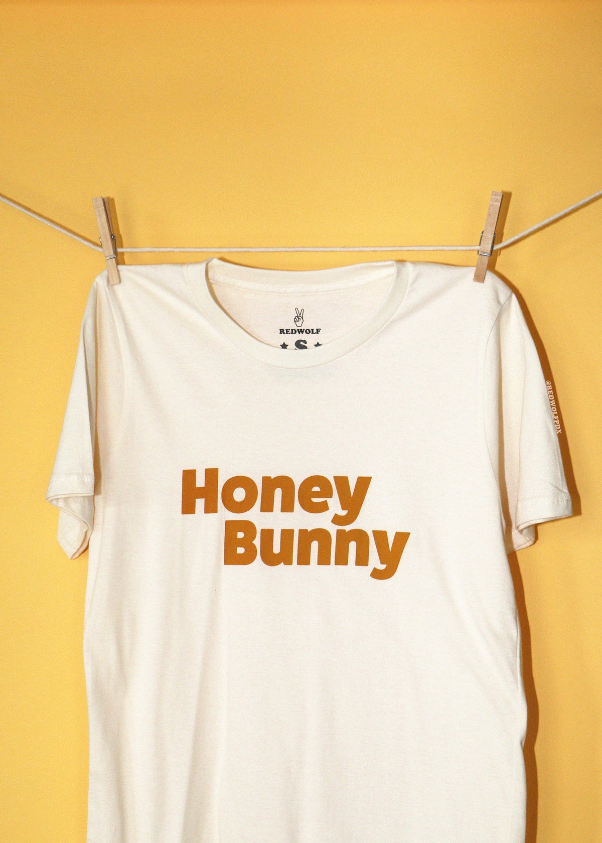 An off-white t-shirt with 'Honey Bunny' printed in orange across the chest, hanging from a clothesline with wooden clothespins against a solid yellow background.