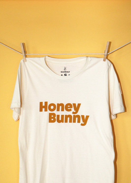 An off-white t-shirt with 'Honey Bunny' printed in orange across the chest, hanging from a clothesline with wooden clothespins against a solid yellow background.