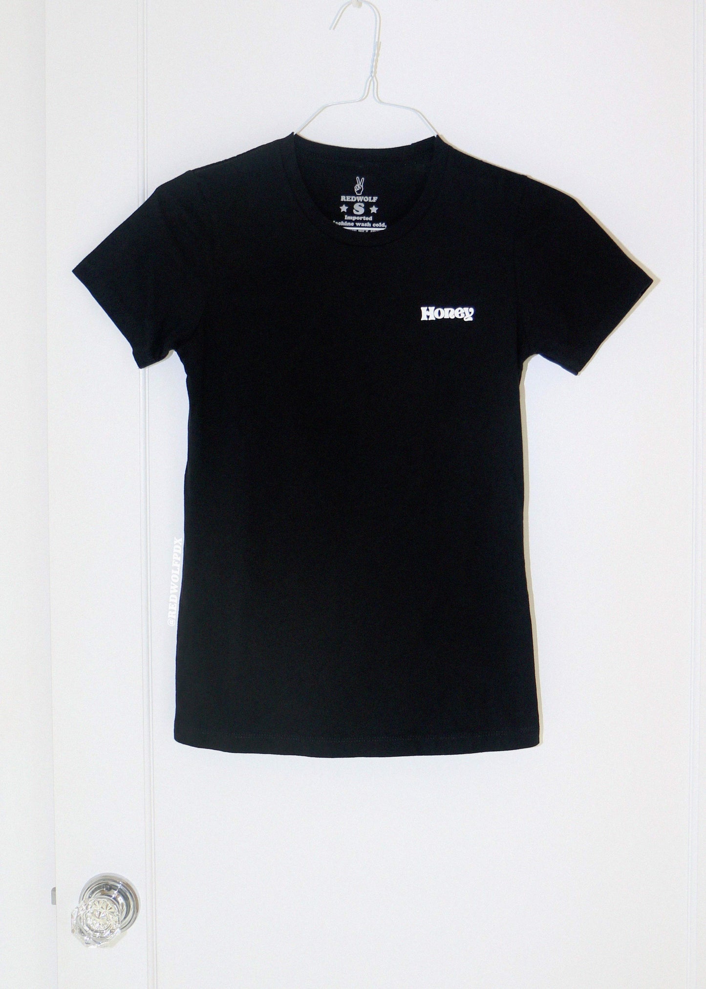 Black t-shirt with  text 'Honey' hanging on on a white door