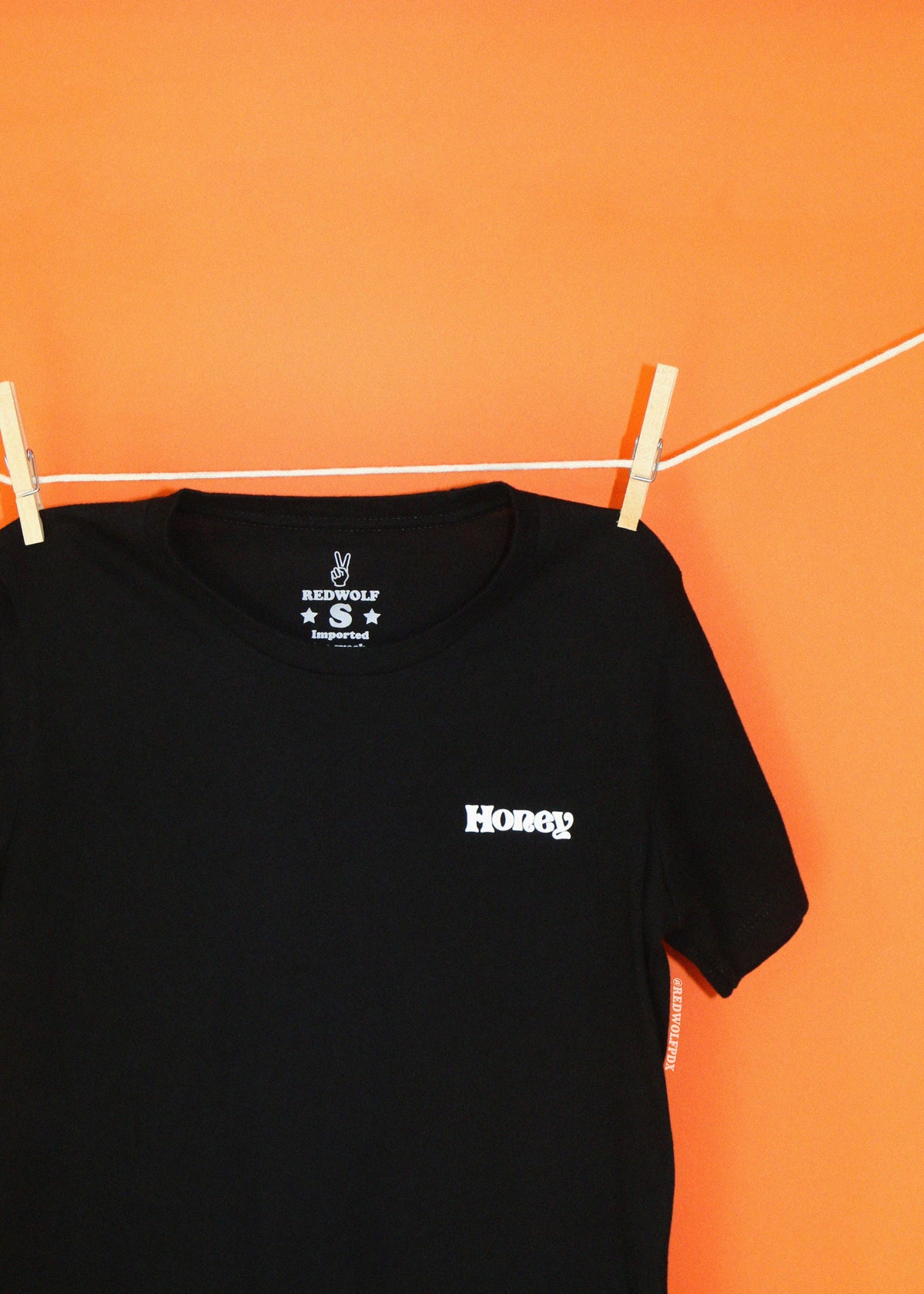 A black short-sleeve t-shirt hanging from a white clothesline with wooden clothespins against a solid orange wall. The shirt features a small white 'Honey' screen printed on the left chest.