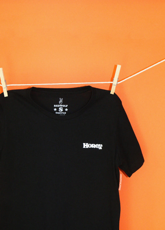 A black short-sleeve t-shirt hanging from a white clothesline with wooden clothespins against a solid orange wall. The shirt features a small white 'Honey' screen printed on the left chest.