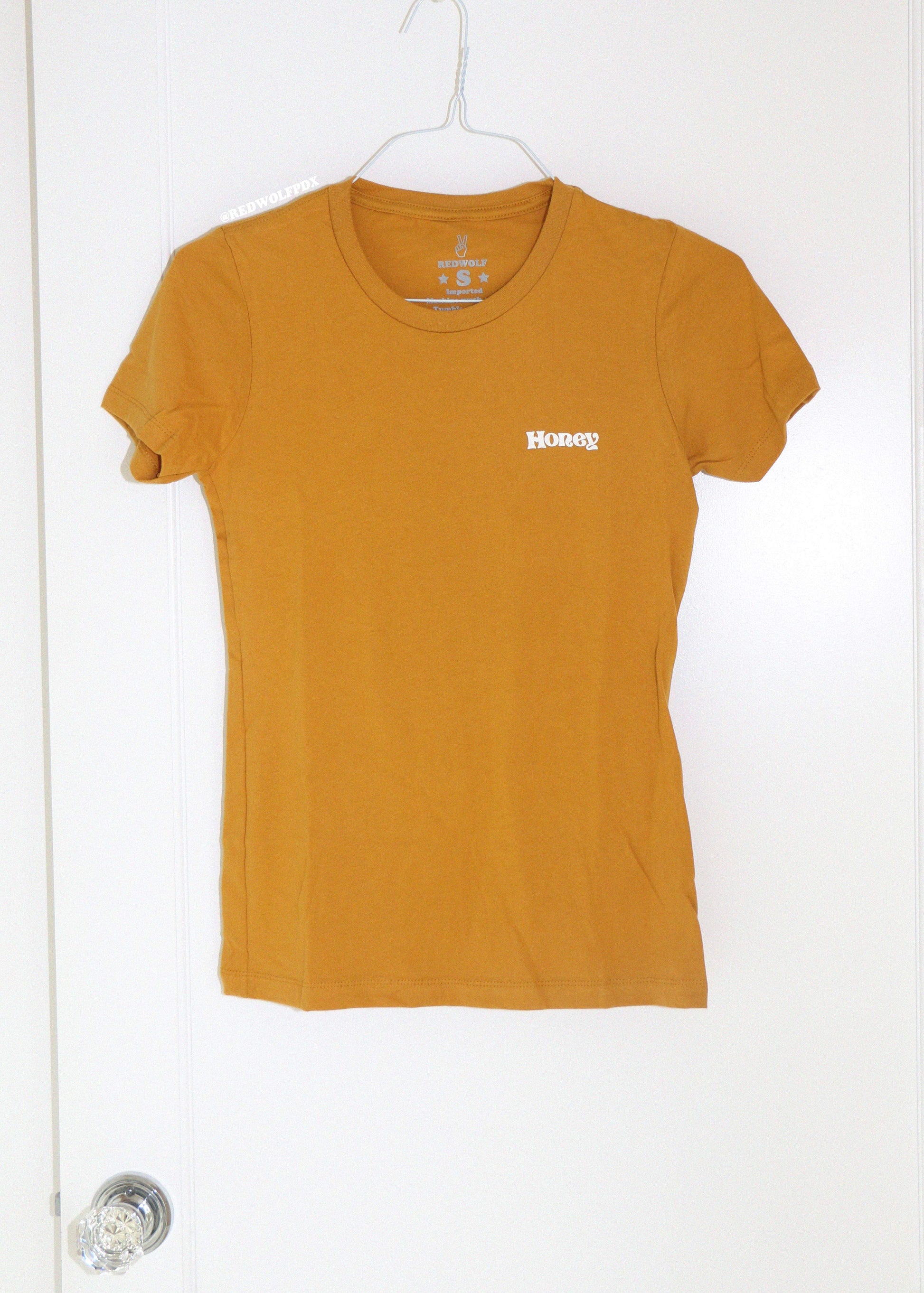 Mustard yellow t-shirt with text 'Honey' on a white hanger against a white background