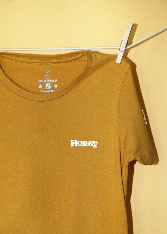 A vintage golden yellow t-shirt clipped to a white clothesline against yellow backdrop. The fitted tee has the text 'Honey' screen printed in white in a 70's font on the left chest.