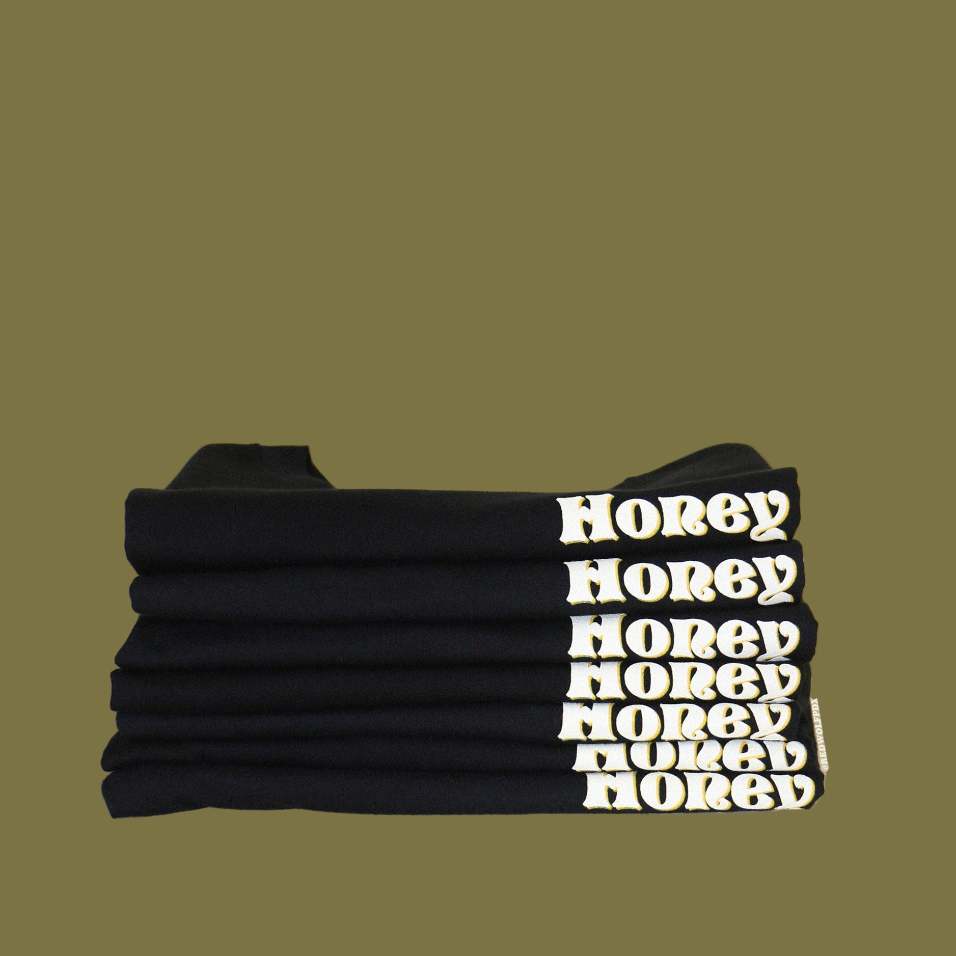 Folded pile of black graphic tees with text 'Honey' in white ink shadowed with yellow ink screen printed on front corner against a beige backdrop