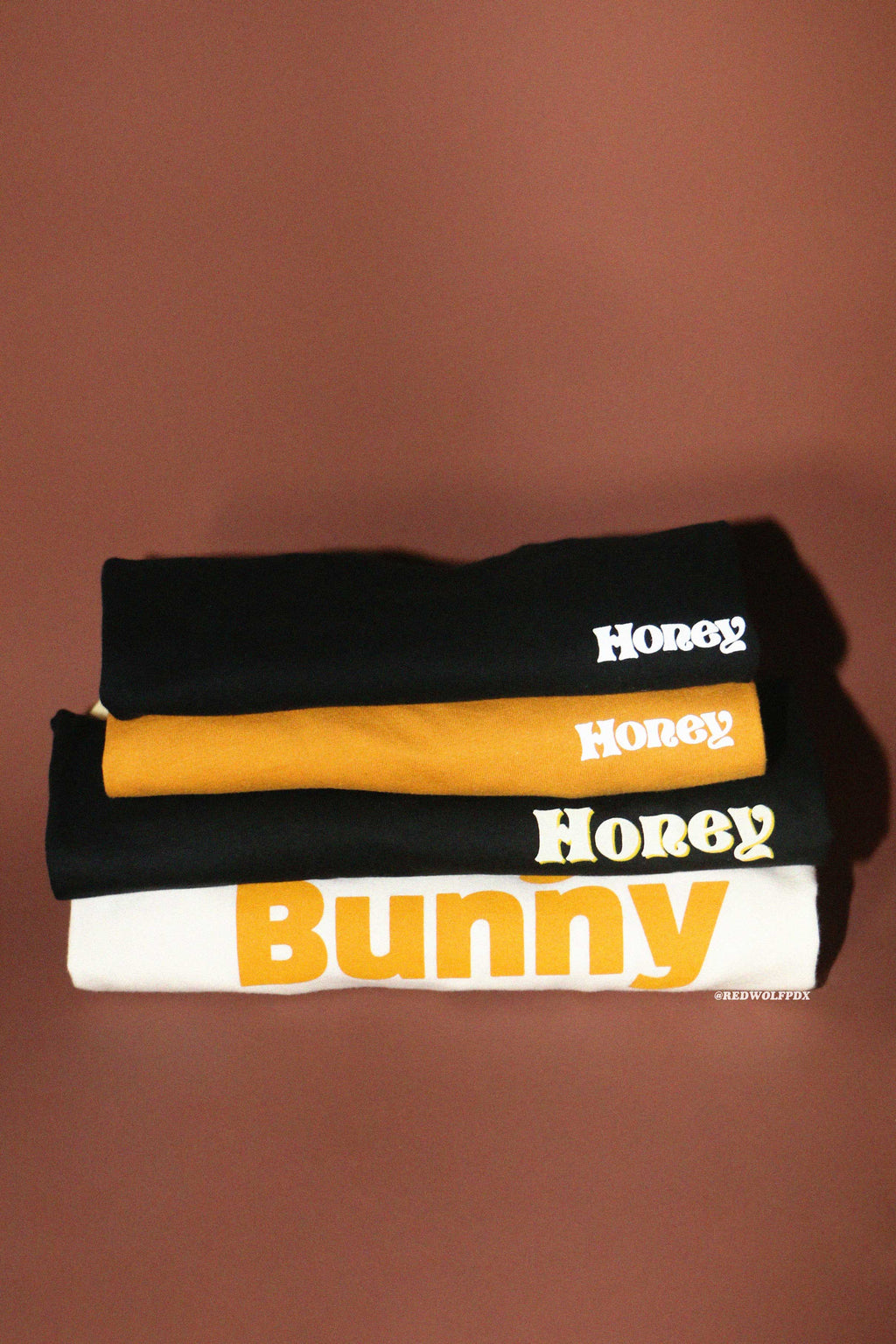 Stack of t-shirts with 'Honey' and 'Honey Bunny' text on a brown background