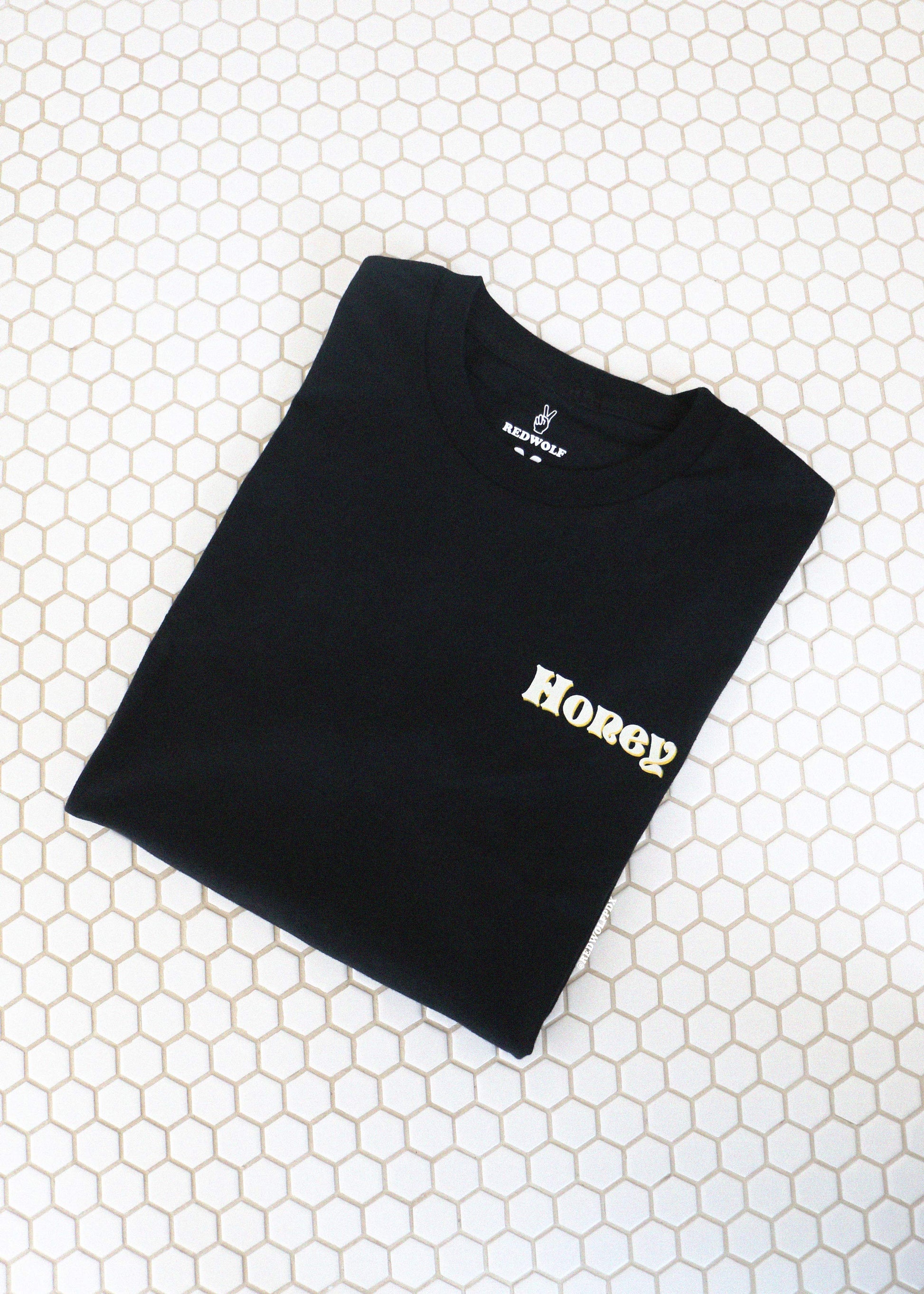 Folded black t-shirt with 'Honey' text on a white tiled floor