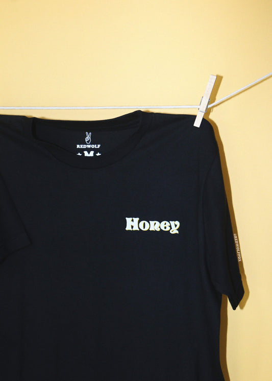 A black short-sleeve t-shirt hanging from a white clothesline with a white plastic clip against a solid pale yellow wall. The shirt has the text 'Honey' printed in a 70's font screen printed in white ink on the left chest.