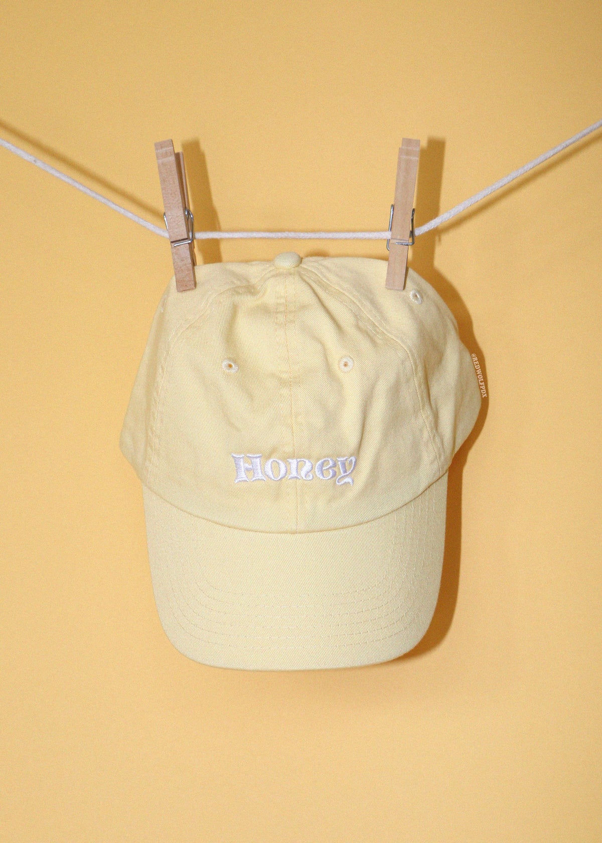 Honey baseball cap in butter yellow with text 'Honey' embroidered in white thread across the front hanging on clothing line against a yellow backdrop.