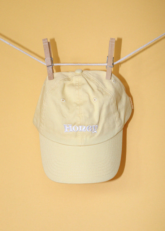 Honey baseball cap in butter yellow with text 'Honey' embroidered in white thread across the front hanging on clothing line against a yellow backdrop.