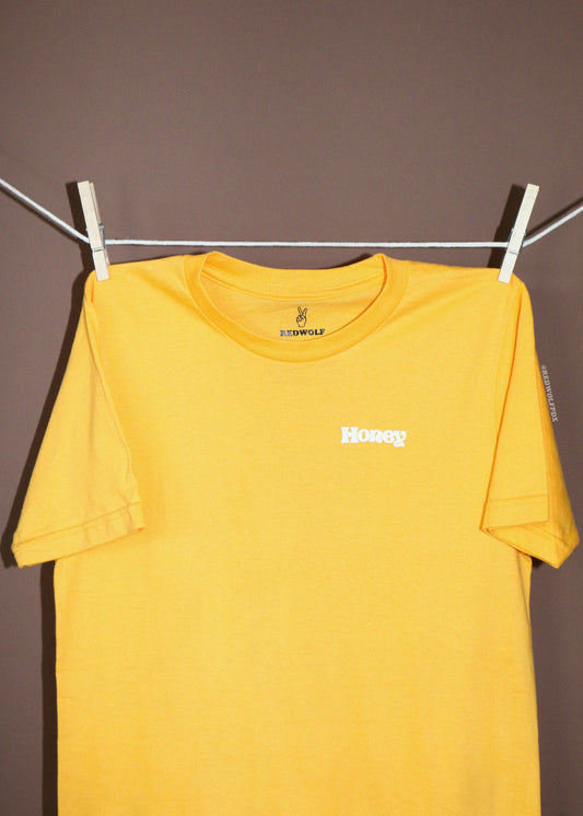 A yellow short-sleeve t-shirt hanging from a white clothesline with white wooden clothespins against a solid dark brown wall. The shirt features a small white 'Honey' graphic screen printed on the left chest.