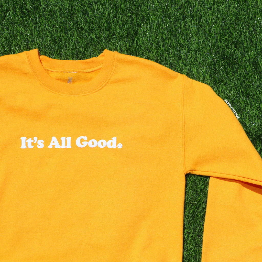 Yellow crewneck sweatshirt with text 'It's all good' laying on grass backdrop