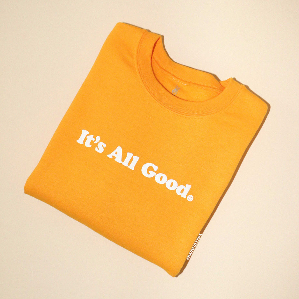 Yellow crewneck sweatshirt with text 'it's all good' folded on beige backdrop