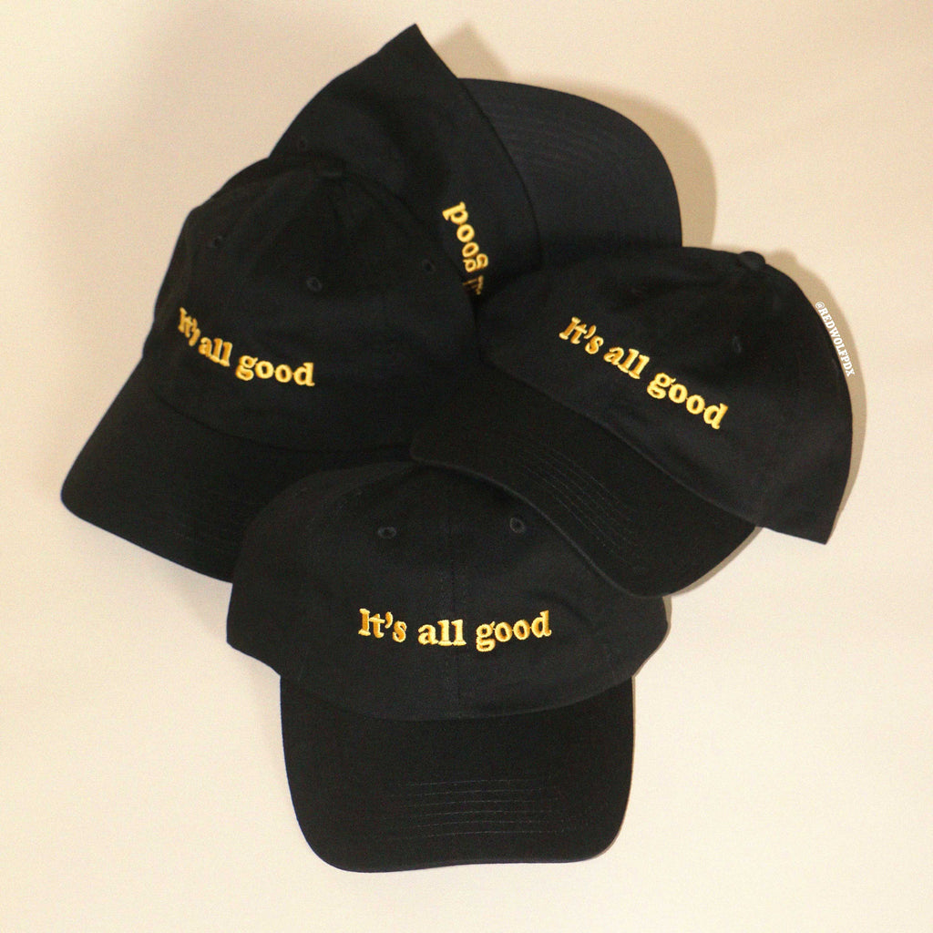 Pile of black baseball caps embroidered with the text 'It's all good' in yellow thread on beige backdrop 