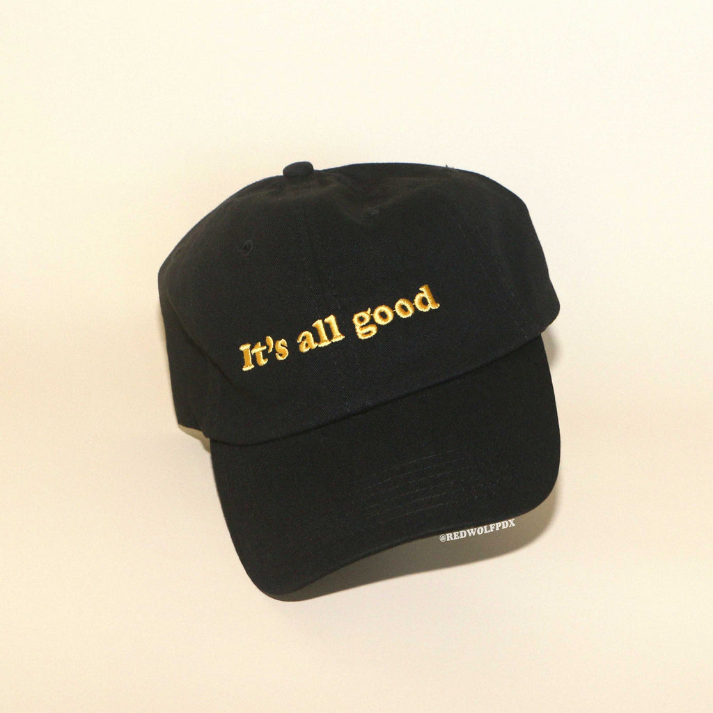 Black baseball cap embroidered with the text 'It's all good' in yellow thread on beige backdrop