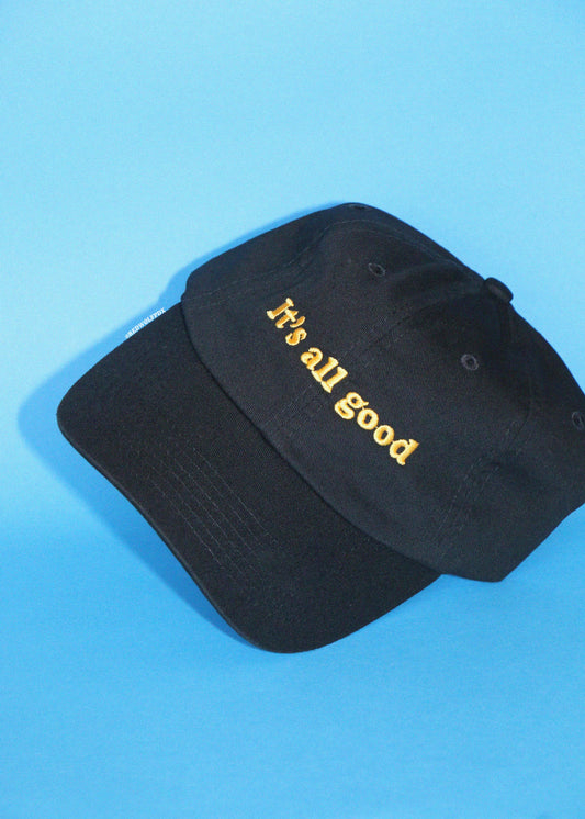 A black baseball cap with 'It's all good' embroidered in yellow on the front, displayed on a solid blue background.