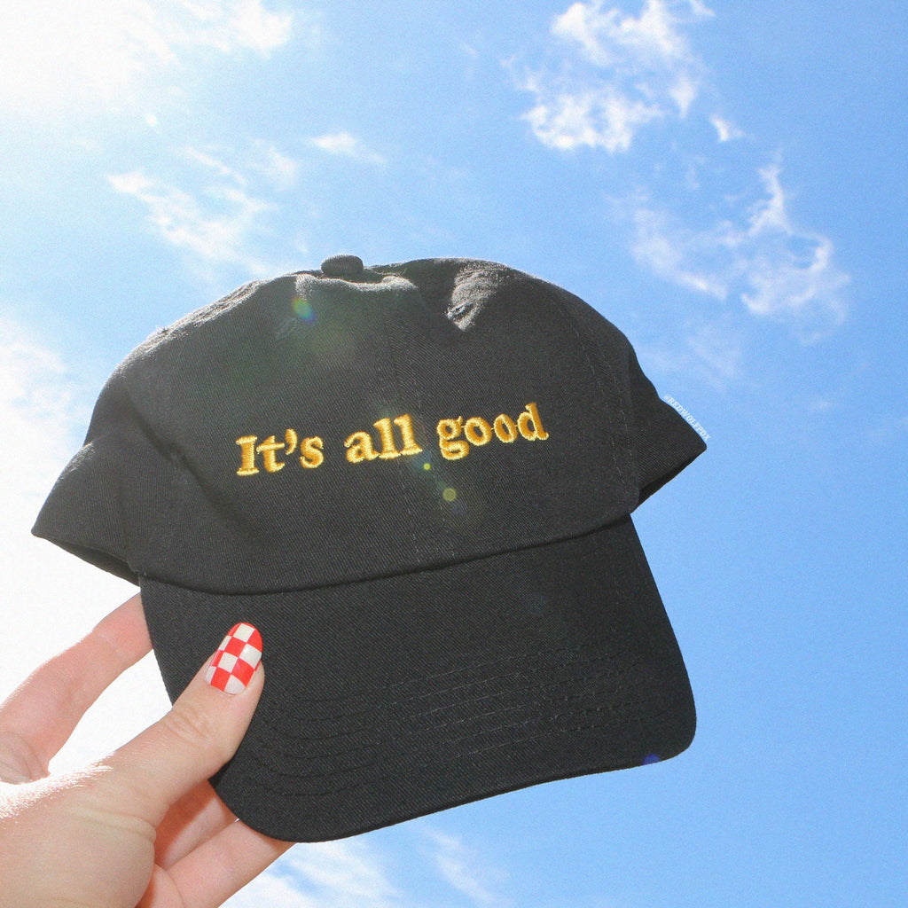 Black baseball cap embroidered with text 'It's all good' in yellow thread being held up to blue sky in background