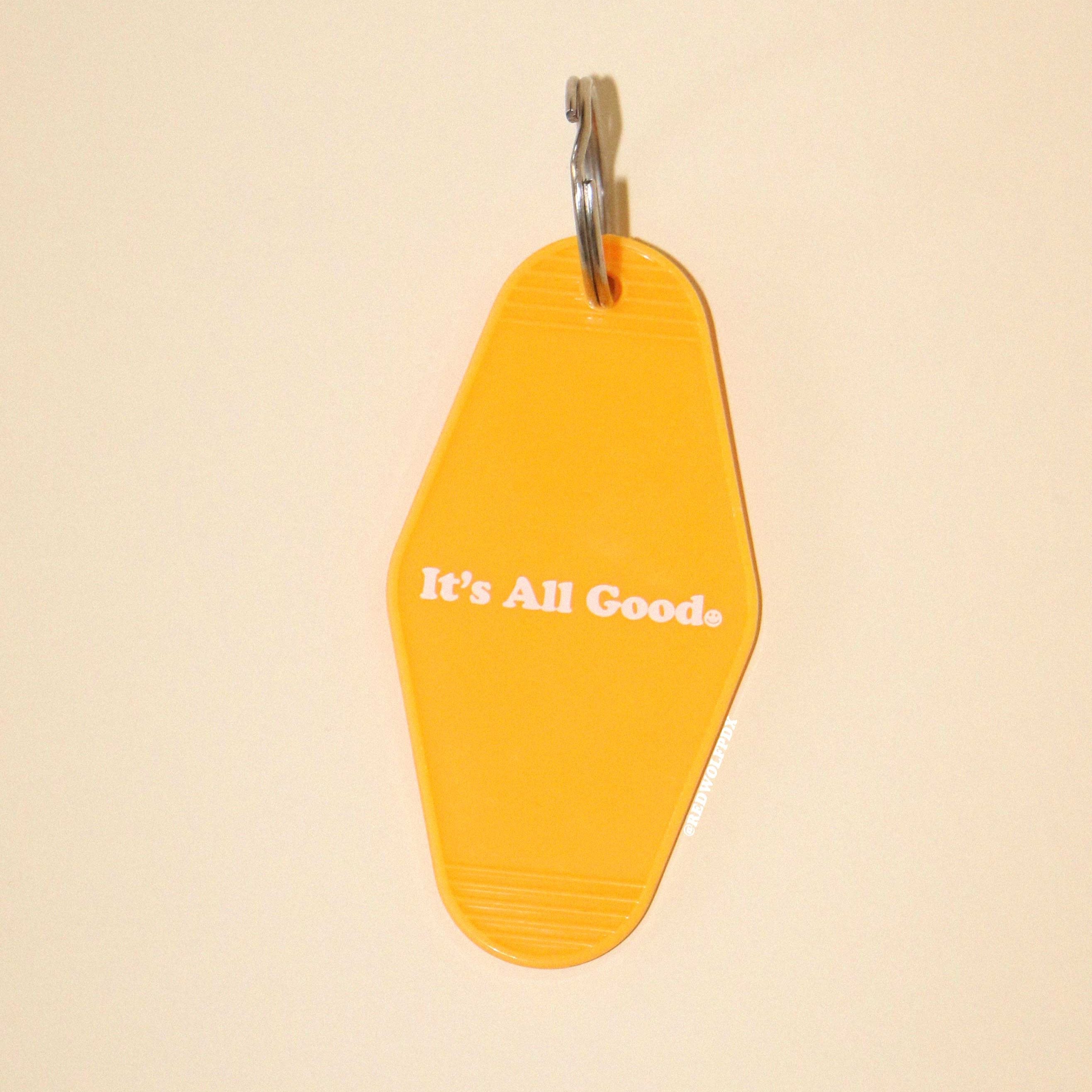 yellow gold retro motel style keychain with text 'It's all good' with smiley face illustration graphic across front against a beige backdrop